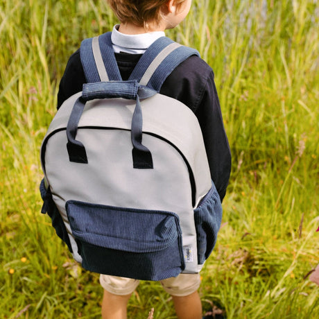 Kind met een lichtgrijze Flashbag Luca rugzak met donkerblauwe voor- en zijvakken tussen het hoge gras.