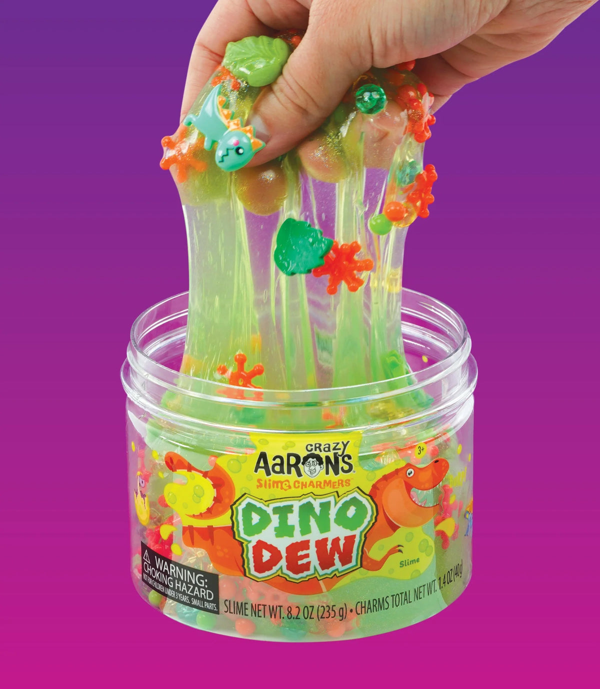 Een hand rekt felgroen Crazy Aaron's Dino Dew slijm met dinosaurus-bedeltjes en kralen uit een open pot.