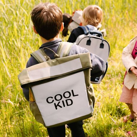 Kind met olijfgroene Flashbag Billie rugzak met zilvergrijs voorvak en tekst 'cool kid' tussen hoog gras.