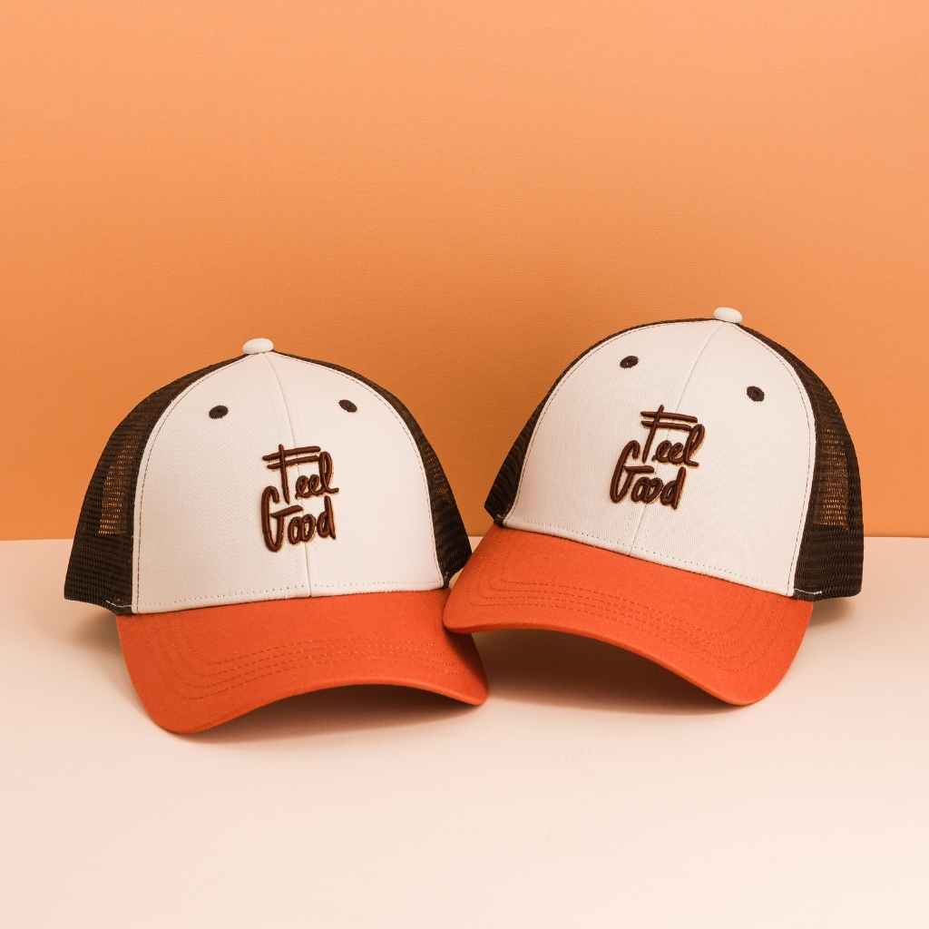 Twee Feel Good-petten met off-white voorkant, bruin borduursel en roestkleurige klep tegen een oranje achtergrond.