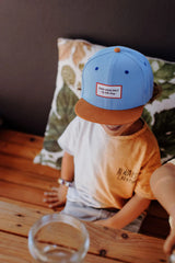 Kind aan een houten tafel met de blauwe Mini Jean pet van Hello Hossy, met de bruine klep naar voren.