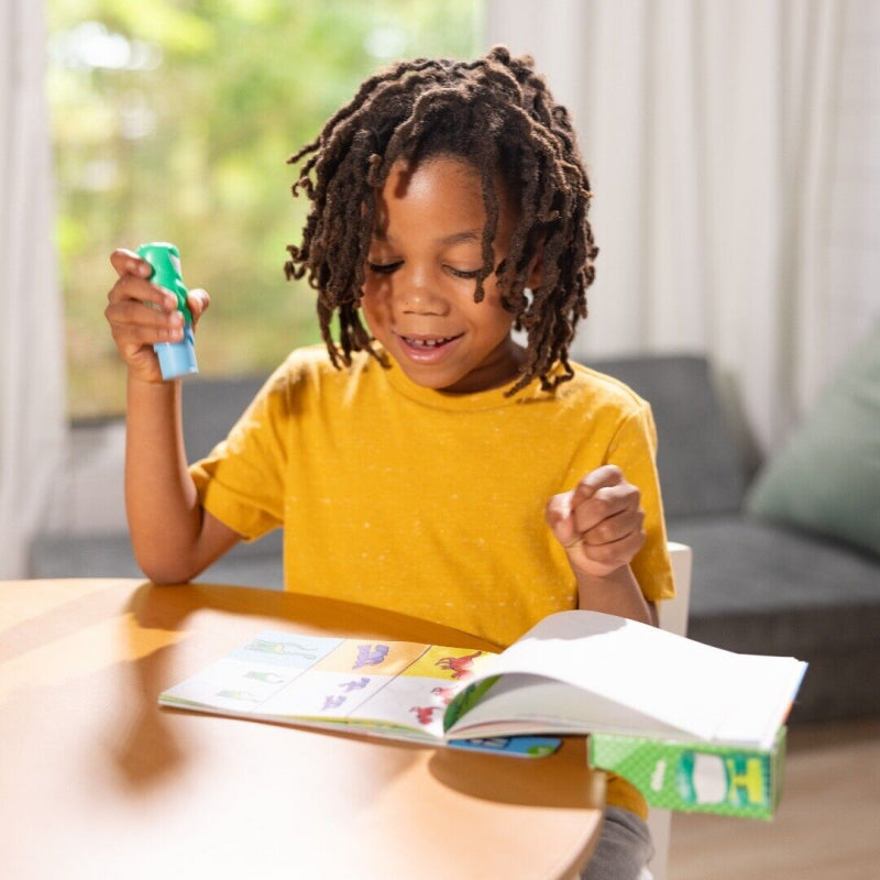 Kind houdt de groene dinosaurus-stickerstempel omhoog bij een open activiteitenboek op tafel.