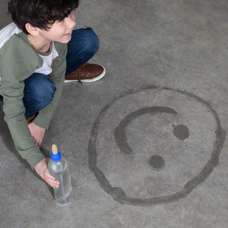 Kind met een waterborstel op een fles naast een met water getekend gezicht op beton.